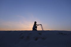 Το όμορφο κορίτσι κάθεται στο σκύψιμο στον αμμώδη λόφο ερήμων στο ηλιοβασίλεμα στοκ εικόνες Το όμορφο κορίτσι κάθεται στο σκύψιμο στον αμμώδη λόφο ερήμων στο ηλιοβασίλεμα στοκ εικόνες