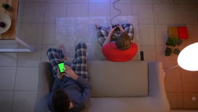 Τοπ πυροβολισμός των φίλων videogame παιχνιδιού πιτζαμάτων με το πηδάλιο και εργασία με το smartphone στο καθιστικό απόθεμα βίντεο Τοπ πυροβολισμός των φίλων videogame παιχνιδιού πιτζαμάτων με το πηδάλιο και εργασία με το smartphone στο καθιστικό απόθεμα βίντεο