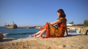 Χαριτωμένη νέα γυναίκα σε ένα θερινά φόρεμα και τα γυαλιά ηλίου που κάθεται από τη δύσκολη ακτή στο υπόβαθρο της γραφικής άποψης φιλμ μικρού μήκους Χαριτωμένη νέα γυναίκα σε ένα θερινά φόρεμα και τα γυαλιά ηλίου που κάθεται από τη δύσκολη ακτή στο υπόβαθρο της γραφικής άποψης φιλμ μικρού μήκους