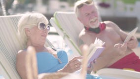 Χαριτωμένο ζεύγος που βρίσκεται στα sunbeds κοντά στη λίμνη Ωριμάστε τη γυναίκα που διαβάζει ένα βιβλίο ενώ ηληκιωμένος που εξετά φιλμ μικρού μήκους Χαριτωμένο ζεύγος που βρίσκεται στα sunbeds κοντά στη λίμνη Ωριμάστε τη γυναίκα που διαβάζει ένα βιβλίο ενώ ηληκιωμένος που εξετά φιλμ μικρού μήκους