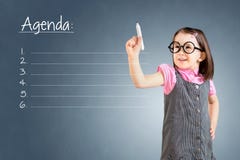 Χαριτωμένο μικρό κορίτσι που φορά το επιχειρησιακό φόρεμα και που γράφει τον κενό κατάλογο ημερήσιων διατάξεων μπλε υπόβαθρο στοκ φωτογραφία με δικαίωμα ελεύθερης χρήσης Χαριτωμένο μικρό κορίτσι που φορά το επιχειρησιακό φόρεμα και που γράφει τον κενό κατάλογο ημερήσιων διατάξεων μπλε υπόβαθρο στοκ φωτογραφία με δικαίωμα ελεύθερης χρήσης