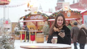 Όμορφη νέα γυναίκα χρησιμοποιώντας ένα smartphone και πίνοντας το καυτό τσάι κατά τη διάρκεια της έκθεσης Χριστουγέννων απόθεμα βίντεο Όμορφη νέα γυναίκα χρησιμοποιώντας ένα smartphone και πίνοντας το καυτό τσάι κατά τη διάρκεια της έκθεσης Χριστουγέννων απόθεμα βίντεο