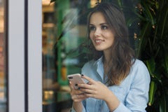 Όμορφη χαριτωμένη καυκάσια νέα γυναίκα στον καφέ, χρησιμοποιώντας το κινητό τηλέφωνο και στεμένος κοντά στο χαμόγελο παραθύρων στοκ φωτογραφία Όμορφη χαριτωμένη καυκάσια νέα γυναίκα στον καφέ, χρησιμοποιώντας το κινητό τηλέφωνο και στεμένος κοντά στο χαμόγελο παραθύρων στοκ φωτογραφία