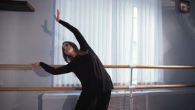 Όμορφος θηλυκός χορευτής μπαλέτου στο μαύρο κοστούμι μεταξιού που στέκεται κοντά στην μπάρα μπαλέτου στην τάξη και που κάνει τις απόθεμα βίντεο Όμορφος θηλυκός χορευτής μπαλέτου στο μαύρο κοστούμι μεταξιού που στέκεται κοντά στην μπάρα μπαλέτου στην τάξη και που κάνει τις απόθεμα βίντεο