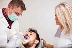 Ώριμος οδοντίατρος και όμορφη νέα γυναίκα βοηθός αρσενικών που χρησιμοποιούν το UV φως, που λευκαίνει τα δόντια ενός νέου αρσενικ στοκ εικόνα με δικαίωμα ελεύθερης χρήσης Ώριμος οδοντίατρος και όμορφη νέα γυναίκα βοηθός αρσενικών που χρησιμοποιούν το UV φως, που λευκαίνει τα δόντια ενός νέου αρσενικ στοκ εικόνα με δικαίωμα ελεύθερης χρήσης
