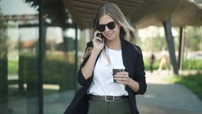 Белокурая женщина в солнечных очках идя с кофе и говоря на smartphone Белокурая женщина в солнечных очках идя с кофе и говоря на smartphone