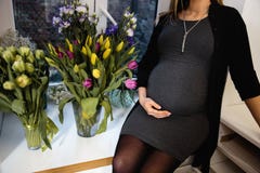 Беременная женщина с тюльпанами цветки держат руки на животе сидит на окне нося черное платье на партии стоковые фотографии rf Беременная женщина с тюльпанами цветки держат руки на животе сидит на окне нося черное платье на партии стоковые фотографии rf