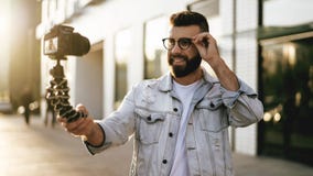 Бородатый мужской блоггер хипстера в ультрамодных стеклах стоя на улице города, держит камеру на блоге треноги и всходов видео- стоковое изображение Бородатый мужской блоггер хипстера в ультрамодных стеклах стоя на улице города, держит камеру на блоге треноги и всходов видео- стоковое изображение
