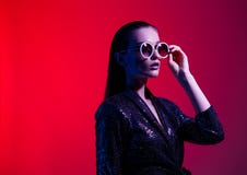 Девушка моды с длинными волосами и круглыми солнечными очками в черном сияющем платье представляет в неоновом свете в студии стоковые фотографии rf Девушка моды с длинными волосами и круглыми солнечными очками в черном сияющем платье представляет в неоновом свете в студии стоковые фотографии rf
