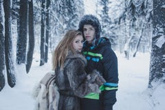Девушка с пропуская волосами в сером пальто и парнем в черных куртке и шляпе на фоне леса зимы стоковая фотография Девушка с пропуская волосами в сером пальто и парнем в черных куртке и шляпе на фоне леса зимы стоковая фотография