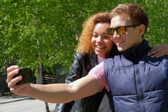 Европейский парень в солнечных очках с темнокожей красивой девушкой, в куртках делает selfie на предпосылке зеленых деревьев стоковое изображение rf Европейский парень в солнечных очках с темнокожей красивой девушкой, в куртках делает selfie на предпосылке зеленых деревьев стоковое изображение rf