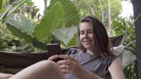 Женщина пишет сообщение на smartphone пока сидящ на гамаке, естественной предпосылке стоковые фотографии rf Женщина пишет сообщение на smartphone пока сидящ на гамаке, естественной предпосылке стоковые фотографии rf