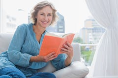 Жизнерадостная белокурая женщина сидя на ее кресле держа книгу Стоковые Фото Жизнерадостная белокурая женщина сидя на ее кресле держа книгу Стоковые Фото