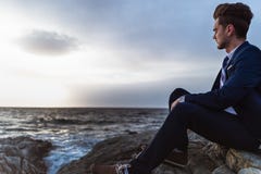 Задумчивый человек в костюме сидит на утесах около моря и думать Молодая ванта стоковые фото Задумчивый человек в костюме сидит на утесах около моря и думать Молодая ванта стоковые фото