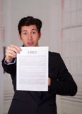 Закройте вверх удивленного молодого бизнесмена нося костюм и держа лист бумаги увольнянного запачканного текста на ем, в Стоковое Изображение RF Закройте вверх удивленного молодого бизнесмена нося костюм и держа лист бумаги увольнянного запачканного текста на ем, в Стоковое Изображение RF