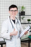 Здравоохранение, профессия и концепция медицины - усмехаясь мужской доктор показывая большие пальцы руки вверх над медицинской пр стоковое изображение rf Здравоохранение, профессия и концепция медицины - усмехаясь мужской доктор показывая большие пальцы руки вверх над медицинской пр стоковое изображение rf