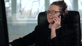 Зрелое брюнет женщины в стеклах работает на компьютере и принимает телефонный звонок с улыбкой сток-видео Зрелое брюнет женщины в стеклах работает на компьютере и принимает телефонный звонок с улыбкой сток-видео