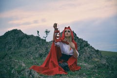 Красивая белокурая девушка с красным плащем на ее голове сидя на утесе стоковое изображение rf Красивая белокурая девушка с красным плащем на ее голове сидя на утесе стоковое изображение rf