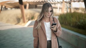 Красивая белокурая женщина в пальто идя и смотря ее smartphone в осени Красивая белокурая женщина в пальто идя и смотря ее smartphone в осени