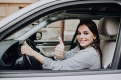 Красивая молодая женщина управляя ее совершенно новым автомобилем и показывая ее большой палец руки вверх стоковое изображение Красивая молодая женщина управляя ее совершенно новым автомобилем и показывая ее большой палец руки вверх стоковое изображение