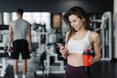 Красивая молодая спортсменка в sportswear и наушниках держит бутылку воды, слушая музыку используя телефон и усмехаться стоковое фото Красивая молодая спортсменка в sportswear и наушниках держит бутылку воды, слушая музыку используя телефон и усмехаться стоковое фото