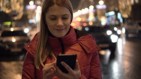 Красивая привлекательная молодая женщина стоя в улице и используя ее smartphone Город освещает автомобили акции видеоматериалы Красивая привлекательная молодая женщина стоя в улице и используя ее smartphone Город освещает автомобили акции видеоматериалы