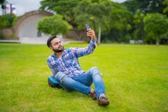 Красивый индийский человек в парке лежа вниз на траве и принимая Selfie с мобильным телефоном стоковая фотография rf Красивый индийский человек в парке лежа вниз на траве и принимая Selfie с мобильным телефоном стоковая фотография rf