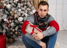 Красивый молодой человек с шарфом сидя с подарками на шотландке около рождественской елки стоковые изображения Красивый молодой человек с шарфом сидя с подарками на шотландке около рождественской елки стоковые изображения