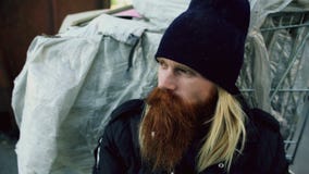 Крупный план молодого бородатого бездомного человека сидя около upet магазинной тележкаи и усиленного самостоятельно в улице стоковые фотографии rf Крупный план молодого бородатого бездомного человека сидя около upet магазинной тележкаи и усиленного самостоятельно в улице стоковые фотографии rf