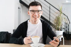 Крытая съемка молодого усмехаясь человека сидя в кофейне Красивый кавказский парень на кафе используя умный телефон стоковое изображение Крытая съемка молодого усмехаясь человека сидя в кофейне Красивый кавказский парень на кафе используя умный телефон стоковое изображение