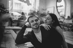Любя девушка держит ее руку на плече ее парня сидя на таблице в уютном романтичном кафе Черный и стоковая фотография Любя девушка держит ее руку на плече ее парня сидя на таблице в уютном романтичном кафе Черный и стоковая фотография