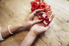 Малая милая присутствующая коробка с смычком в красивых руках женщины Фокус на смычке Цветки красных роз позади на деревянном сто стоковое изображение Малая милая присутствующая коробка с смычком в красивых руках женщины Фокус на смычке Цветки красных роз позади на деревянном сто стоковое изображение