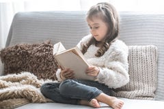 Маленькая девочка читая книгу на удобной софе, красивых эмоциях стоковая фотография rf Маленькая девочка читая книгу на удобной софе, красивых эмоциях стоковая фотография rf