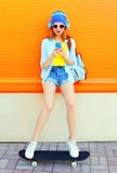 Мода довольно холодная девушка слушает к музыке и использование smartphone сидит на скейтборде над красочным апельсином Стоковые Фотографии RF Мода довольно холодная девушка слушает к музыке и использование smartphone сидит на скейтборде над красочным апельсином Стоковые Фотографии RF
