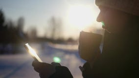 Молодая красивая девушка выпивая горячий кофе в зиме в природе, держа телефон в руке, управляет пальцем через акции видеоматериалы Молодая красивая девушка выпивая горячий кофе в зиме в природе, держа телефон в руке, управляет пальцем через акции видеоматериалы