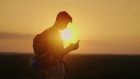 Молодой туристский человек с рюкзаком использует мобильный телефон na górze горы В лучах заходящего солнца, апельсин сток-видео Молодой туристский человек с рюкзаком использует мобильный телефон na górze горы В лучах заходящего солнца, апельсин сток-видео