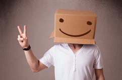 Молодой человек показывать с картонной коробкой на его голове с smiley Стоковые Изображения RF Молодой человек показывать с картонной коробкой на его голове с smiley Стоковые Изображения RF