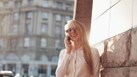 Молодая бизнес-леди стоя близко офисное здание на солнечных улицах города и говоря на передвижном сотовом телефоне видеоматериал Молодая бизнес-леди стоя близко офисное здание на солнечных улицах города и говоря на передвижном сотовом телефоне видеоматериал