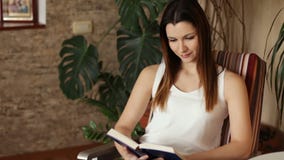Молодая красивая девушка читая книгу сидя в стуле Привлекательная женщина усмехаясь пока читающ книгу взволнованности положительн сток-видео Молодая красивая девушка читая книгу сидя в стуле Привлекательная женщина усмехаясь пока читающ книгу взволнованности положительн сток-видео