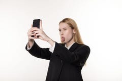 Молодая привлекательная женщина в темном костюме принимает фото используя мобильный телефон, закрывает один глаз и язык показыват стоковое изображение rf Молодая привлекательная женщина в темном костюме принимает фото используя мобильный телефон, закрывает один глаз и язык показыват стоковое изображение rf