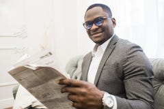 Молодой Афро-американский бизнесмен в сером костюме читая газету пока сидящ на софе стоковое фото rf Молодой Афро-американский бизнесмен в сером костюме читая газету пока сидящ на софе стоковое фото rf
