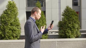 Молодой бизнесмен идя вниз по улице и агрессивно водит обсуждение на видео- звонке Молодой бизнесмен идя вниз по улице и агрессивно водит обсуждение на видео- звонке