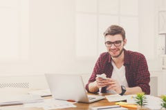 Молодой счастливый бизнесмен в офисе используя сотовый телефон компьтер-книжкой Стоковые Изображения Молодой счастливый бизнесмен в офисе используя сотовый телефон компьтер-книжкой Стоковые Изображения