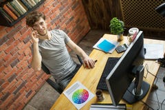 Молодой усмехаясь цифровой дизайнер художника говоря смартфоном с его клиентом на его рабочем месте в офисе стиля просторной квар стоковое фото Молодой усмехаясь цифровой дизайнер художника говоря смартфоном с его клиентом на его рабочем месте в офисе стиля просторной квар стоковое фото
