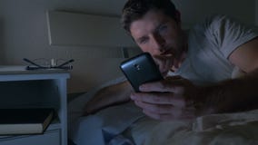 Молодой человек в кресле кровати дома поздно на ноче используя мобильный телефон в нижнем свете ослабил в технике связи Молодой человек в кресле кровати дома поздно на ноче используя мобильный телефон в нижнем свете ослабил в технике связи