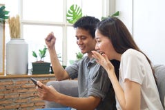 Молодые пары сидя на софе наблюдают мобильный телефон и чувствуют surprise&happy стоковые изображения rf Молодые пары сидя на софе наблюдают мобильный телефон и чувствуют surprise&happy стоковые изображения rf