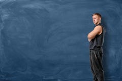 Мышечный молодой человек стоит его пересеченные оружия и смотрит вперед на голубой предпосылке доски стоковое фото Мышечный молодой человек стоит его пересеченные оружия и смотрит вперед на голубой предпосылке доски стоковое фото