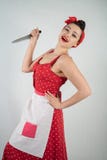 Опасный штырь вверх по домохозяйке девушки в красном винтажном платье точки польки стоит с огромным кухонным ножом в ее руках и с стоковое фото rf Опасный штырь вверх по домохозяйке девушки в красном винтажном платье точки польки стоит с огромным кухонным ножом в ее руках и с стоковое фото rf