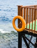 Оранжевая lifebuoy смертная казнь через повешение на коричневой деревянной пристани против сини стоковое изображение Оранжевая lifebuoy смертная казнь через повешение на коричневой деревянной пристани против сини стоковое изображение
