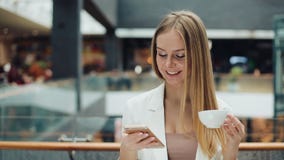 Очаровательная молодая женщина держит smartphone в одной руке и чашке кофе в другом усаживании в кафе видеоматериал Очаровательная молодая женщина держит smartphone в одной руке и чашке кофе в другом усаживании в кафе видеоматериал